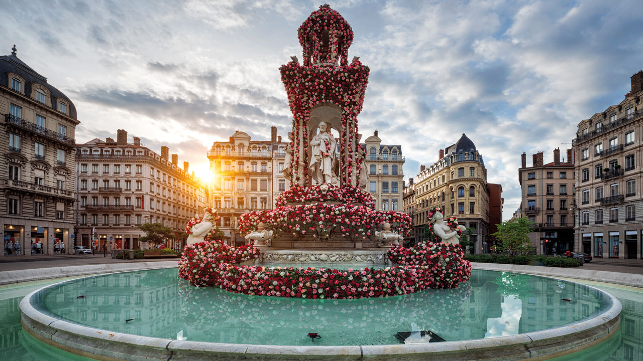 Ein prachtvoller Brunnen in Lyon, welcher mit Blumen geschmückt ist und in Mitten eines Platzes liegt. Die Sonne scheint zwischen Häusern hindurch.