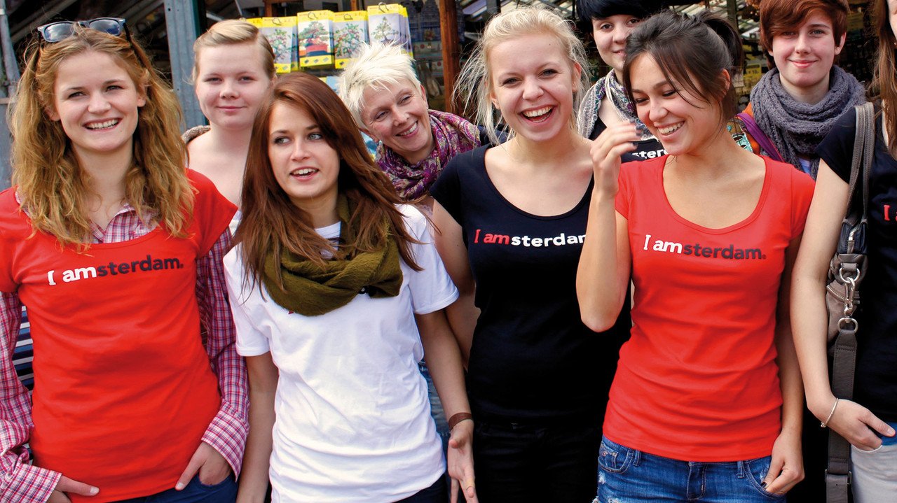 Klassenfahrten Niederlande Eine Gruppe Schülerinnen mit ihrer Lehrerin auf Klassenfahrt in Amsterdam. Alle lachen und tragen rote und schwarze Shirts mit der Aufschrift I amsterdam. Amsteer